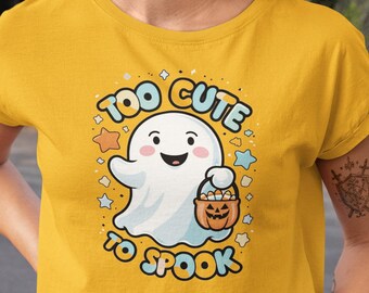Puede incluir: Top corto amarillo mostaza con un diseño de Halloween. El diseño presenta un fantasma blanco sonriente sosteniendo un cubo de calabaza, rodeado de estrellas y el texto "Too Cute To Spook."