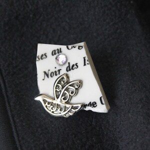 May include: A silver dove brooch with intricate openwork design, set on a piece of paper with French text and a sparkling rhinestone. The brooch is attached to a black fabric.