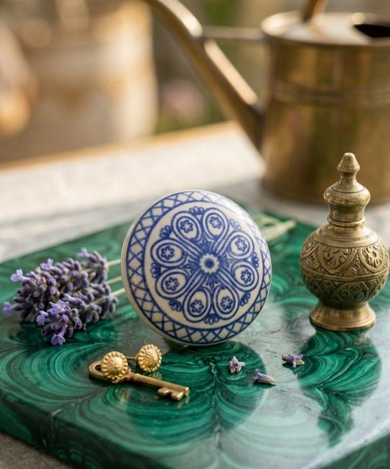 Op de afbeelding: Een decoratieve keramische kastknop met een blauw en wit bloemmotief. Een kleine messing sleutel en oorbellen liggen naast lavendel op een groen oppervlak. Een kleine messing parfumfles en een gieter staan op de achtergrond.