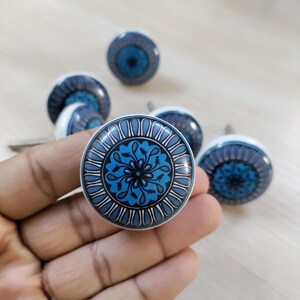 May include: Set of six ceramic cabinet knobs. Each knob features a white base and a blue floral design with a dark blue and black center. The knobs are round and have a metal screw for installation.