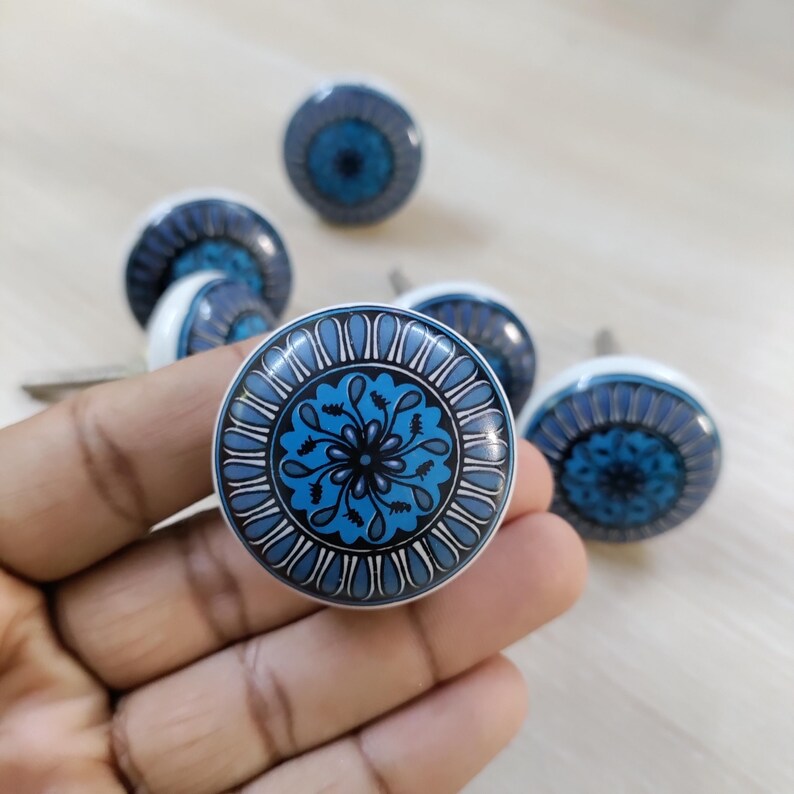 May include: Set of six ceramic cabinet knobs. Each knob features a white base and a blue floral design with a dark blue and black center. The knobs are round and have a metal screw for installation.