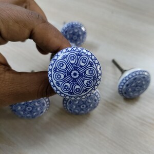 May include: A collection of blue and white ceramic cabinet knobs. Each knob features a detailed, symmetrical design in blue against a white background. The knobs are round with a metal stem for attachment. The design is reminiscent of traditional pottery.