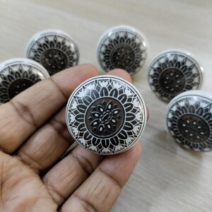 May include: A collection of six white ceramic cabinet knobs with a black floral design. The knobs are round with a central dark brown circle and intricate black patterns. The knobs are shown on a light wood surface.