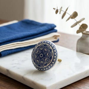 Op de afbeelding: Witte keramische kastknop met een blauw bloemmotief. De knop heeft een messing steel en staat op een wit marmeren oppervlak. Blauwe gevouwen linnen op de achtergrond.