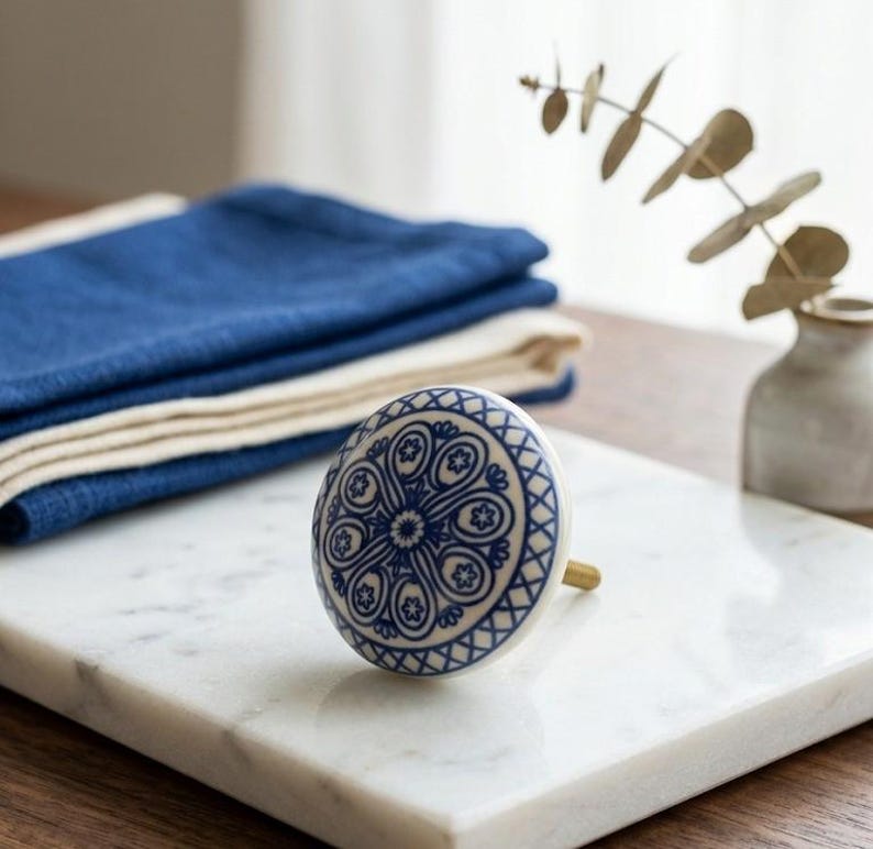 Op de afbeelding: Witte keramische kastknop met een blauw bloemmotief. De knop heeft een messing steel en staat op een wit marmeren oppervlak. Blauwe gevouwen linnen op de achtergrond.