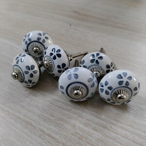 May include: Set of six white ceramic cabinet knobs with a floral pattern in blue and gray. Each knob has a silver-colored metal base and screw. The knobs are arranged on a light wood-grain surface.