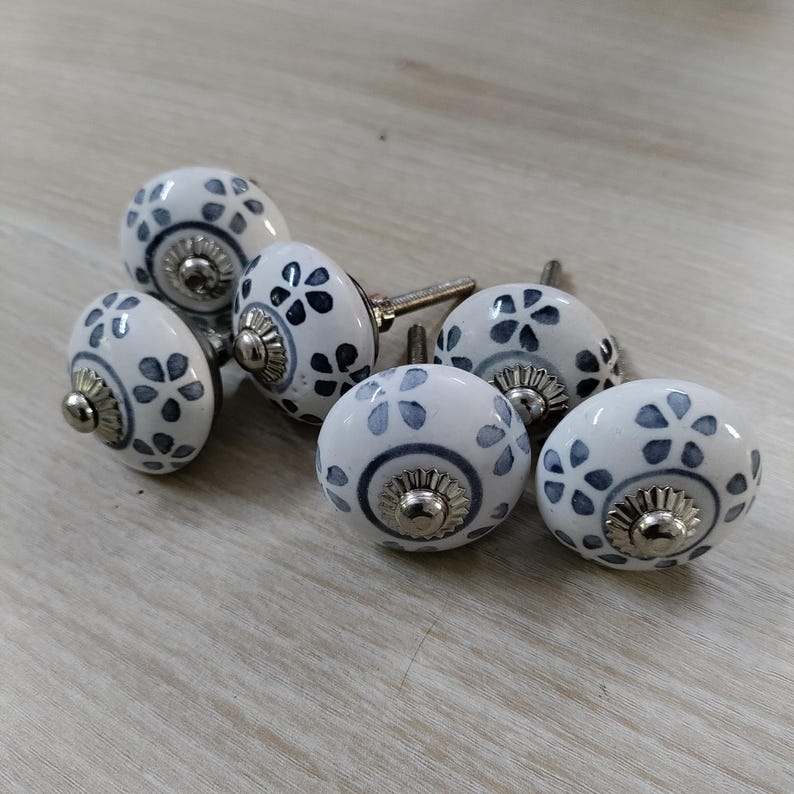 May include: Set of six white ceramic cabinet knobs with a floral pattern in blue and gray. Each knob has a silver-colored metal base and screw. The knobs are arranged on a light wood-grain surface.