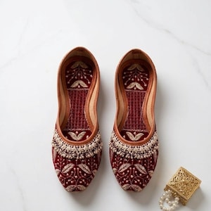 May include: A pair of burgundy velvet slip-on shoes with intricate floral embroidery and a beaded trim. The shoes have a leather lining and a decorative gold box with a pearl necklace is next to them.