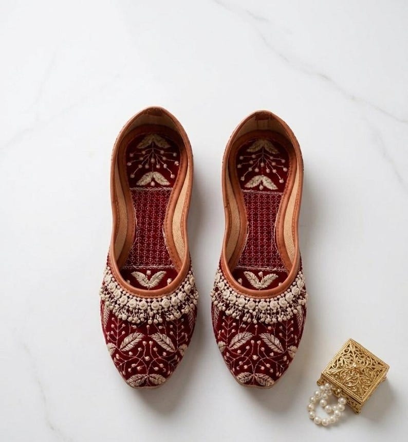 May include: A pair of burgundy velvet slip-on shoes with intricate floral embroidery and a beaded trim. The shoes have a leather lining and a decorative gold box with a pearl necklace is next to them.