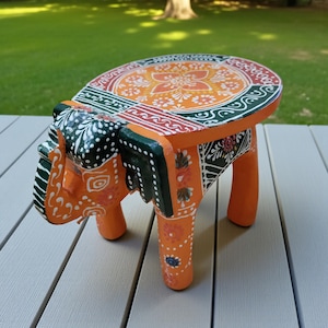Puede incluir: Taburete de elefante de madera naranja pintado a mano con un diseño floral decorativo en la parte superior. El taburete presenta detalles en verde, rojo y blanco. Las patas son naranjas con motivos florales.
