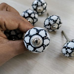 May include: A collection of white ceramic cabinet knobs with a black floral design. Each knob features a silver-colored metal base and a decorative pattern. The knobs are designed for furniture and home decor.