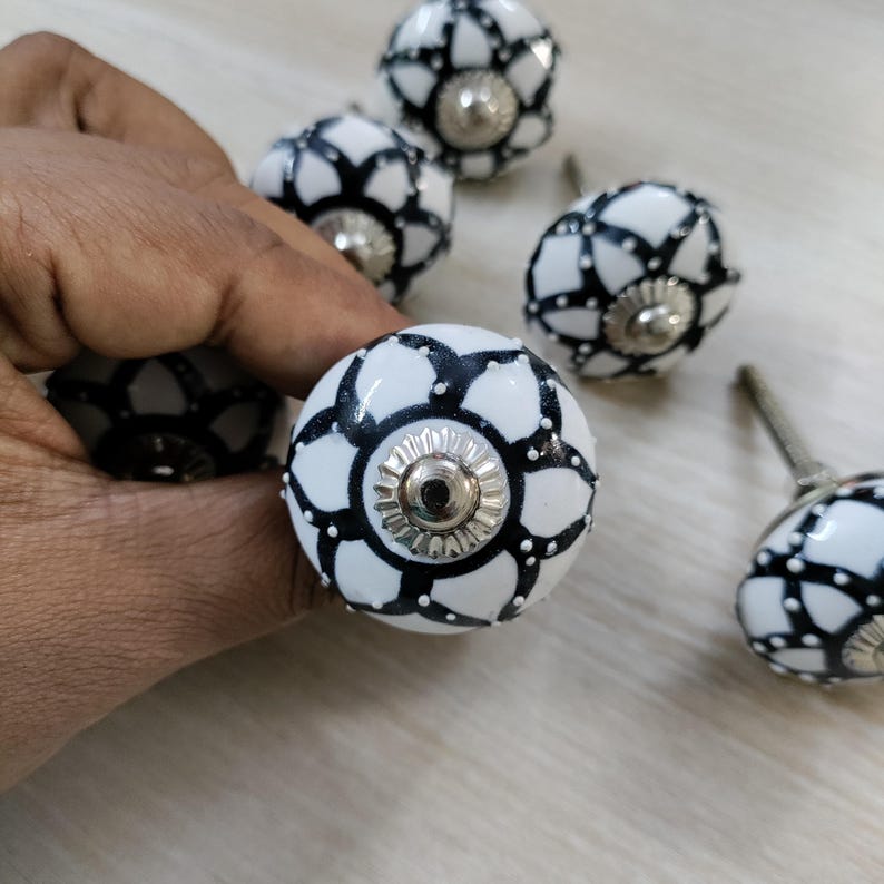 May include: A collection of white ceramic cabinet knobs with a black floral design. Each knob features a silver-colored metal base and a decorative pattern. The knobs are designed for furniture and home decor.