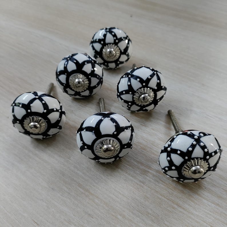 May include: Six white and black decorative cabinet knobs with a floral design. Each knob has a silver-colored base and a screw for installation. The knobs are arranged on a light wood surface.