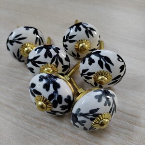 May include: Set of six white ceramic cabinet knobs with black leaf patterns. Each knob has a gold-colored base and screw. The knobs are arranged on a light wood surface, offering a decorative hardware option.