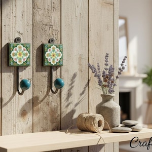 Könnte beinhalten: Zwei dekorative Wandhaken mit quadratischen grünen Fliesen mit floralen Mustern. Jeder Haken hat eine türkisfarbene Kugel am Ende. Die Haken sind an einer hellen Holzwand befestigt. Darunter befindet sich ein Regal mit einer Vase, Bindfaden und Steinen.