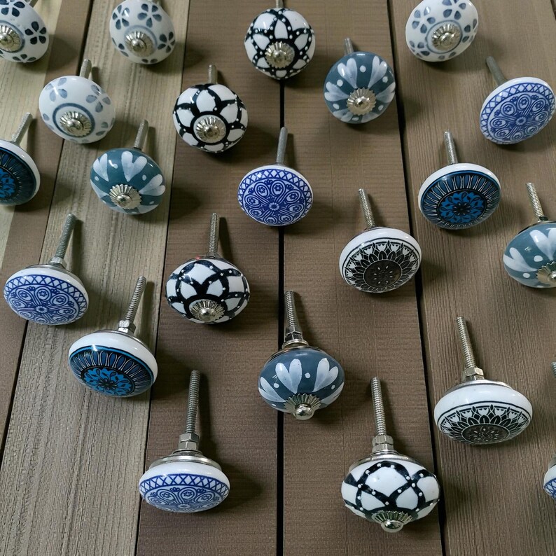 May include: A collection of decorative ceramic cabinet knobs in various designs. The knobs feature patterns in white, blue, and black, with silver-toned metal stems. The knobs are arranged on a wooden surface, showcasing their diverse styles.