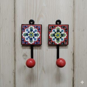 Könnte beinhalten: Zwei dekorative Wandhaken. Jeder Haken hat eine quadratische, rote Fliese mit einem Blumenmuster in Blau, Gelb und Weiß. Sie haben schwarze Metallrahmen und -stiele mit einer roten Kugel am Ende.
