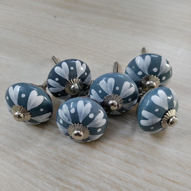 May include: A set of six decorative cabinet knobs. Each knob is a rounded shape, painted in a muted blue-gray color with a white floral design. The knobs have silver-colored metal hardware.