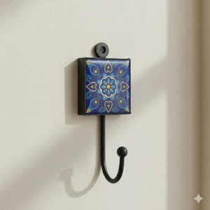 May include: A wall-mounted hook featuring a square tile with a blue and yellow floral pattern. The tile is framed in black metal, with a black hook below. The hook is attached to a white wall.