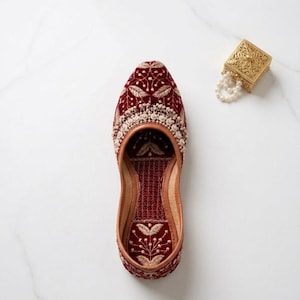 May include: A maroon velvet shoe with intricate gold embroidery and a cream-colored beaded trim. The shoe features a floral design and a leather sole. A small gold box with a pearl necklace is in the background.