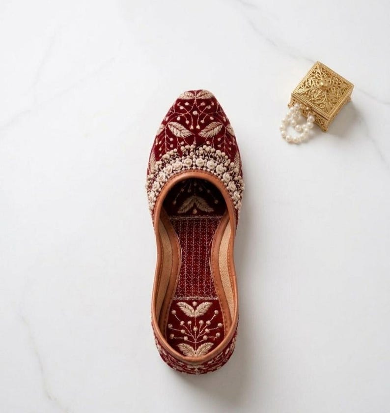 May include: A maroon velvet shoe with intricate gold embroidery and a cream-colored beaded trim. The shoe features a floral design and a leather sole. A small gold box with a pearl necklace is in the background.