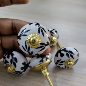 May include: Set of five white ceramic cabinet knobs with black leaf patterns and gold-colored metal accents. Each knob features a gold-colored flower-shaped center. The knobs are designed for furniture and home decor.