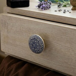 Op de afbeelding: Close-up van een lichtgekleurde houten lade met een decoratieve blauw-witte keramische knop. De knop heeft een cirkelvormig ontwerp. De lade maakt deel uit van een groter meubelstuk, met lavendel en een kleine gouden vaas.