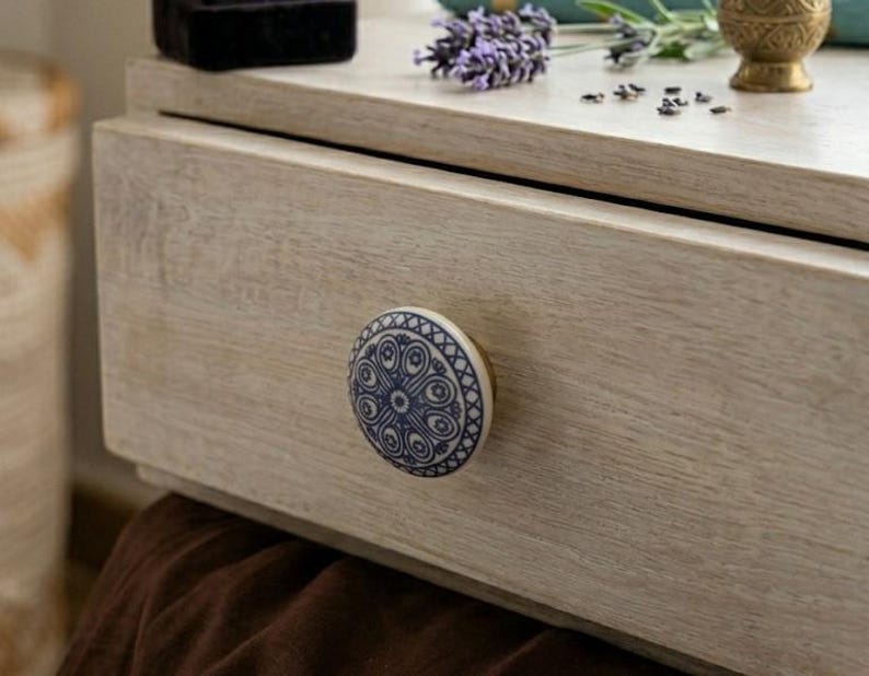 Op de afbeelding: Close-up van een lichtgekleurde houten lade met een decoratieve blauw-witte keramische knop. De knop heeft een cirkelvormig ontwerp. De lade maakt deel uit van een groter meubelstuk, met lavendel en een kleine gouden vaas.