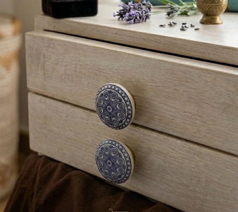Op de afbeelding: Een lichtgekleurde houten dressoir met twee lades, elk versierd met een ronde, wit en blauw patroonknop. De knoppen hebben een decoratief ontwerp. De dressoir is bedekt met decoratieve items.