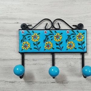 Könnte beinhalten: Ein dekorativer Wandhaken mit einer türkisfarbenen Keramikfliese mit gelben Blumenmustern. Der Haken hat drei hellblaue kugelförmige Haken und einen schwarzen Metallrahmen mit dekorativer Oberseite.