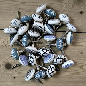 Op de afbeelding: Een verzameling decoratieve keramische kastknoppen in diverse designs. De knoppen hebben witte en blauwe patronen, waaronder bloemen, geometrische en abstracte motieven. Elke knop heeft een metalen schroef voor bevestiging. Geplaatst op een houten oppervlak.