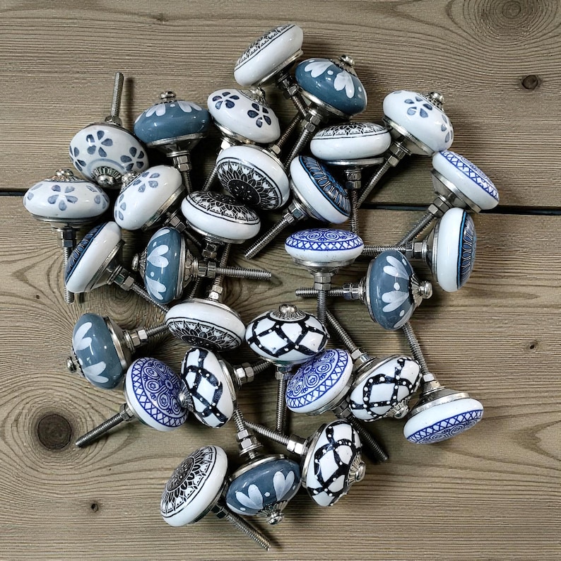 May include: A collection of decorative ceramic cabinet knobs in various designs. The knobs feature white and blue patterns, including floral, geometric, and abstract motifs. Each knob has a metal screw for installation. The knobs are arranged on a wooden surface.