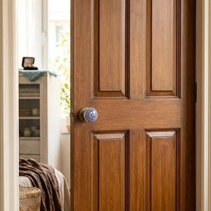 Op de afbeelding: Een houten deur met een warme, bruine afwerking en een decoratieve blauw-witte deurknop. De deur heeft een klassiek paneelontwerp en staat gedeeltelijk open, waardoor een glimp van een slaapkamerinterieur te zien is.