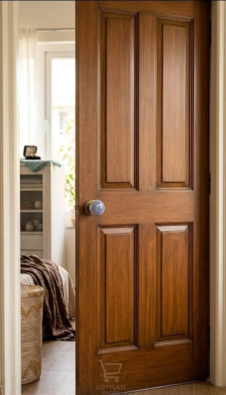 Op de afbeelding: Een houten deur met een warme, bruine afwerking en een decoratieve blauw-witte deurknop. De deur heeft een klassiek paneelontwerp en staat gedeeltelijk open, waardoor een glimp van een slaapkamerinterieur te zien is.