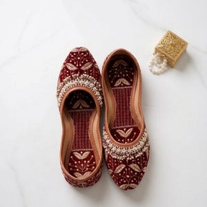 May include: A pair of burgundy velvet embroidered shoes with intricate floral patterns and cream-colored beadwork. The shoes have a leather trim and a matching burgundy interior. A small gold box and a string of pearls are also in the image.