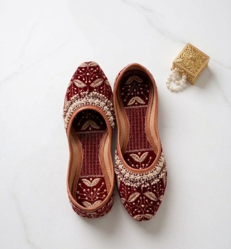 May include: A pair of burgundy velvet embroidered shoes with intricate floral patterns and cream-colored beadwork. The shoes have a leather trim and a matching burgundy interior. A small gold box and a string of pearls are also in the image.