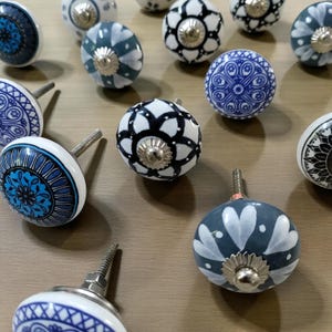 May include: A collection of decorative ceramic cabinet knobs in various designs. The knobs feature patterns in blue, white, and black, with silver-toned metal hardware. The designs include floral, geometric, and abstract motifs, suitable for furniture or home decor.