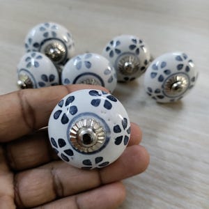 May include: A collection of white ceramic cabinet knobs with a floral design in navy blue. Each knob has a silver-colored metal base. The knobs are round and have a glossy finish, suitable for furniture or decor.