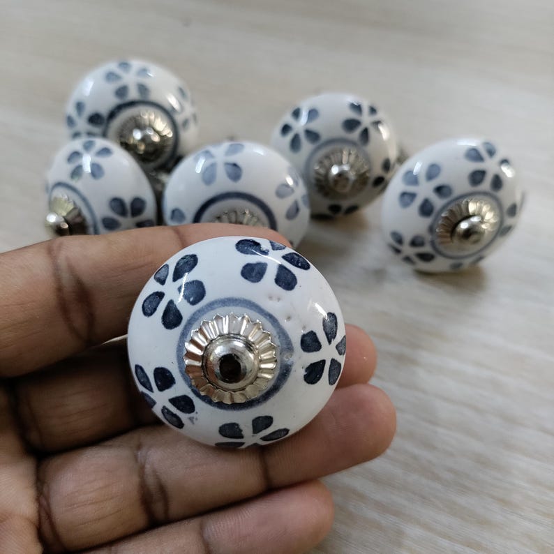 May include: A collection of white ceramic cabinet knobs with a floral design in navy blue. Each knob has a silver-colored metal base. The knobs are round and have a glossy finish, suitable for furniture or decor.