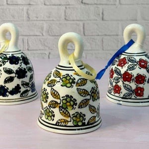 May include: Three ceramic bells with floral patterns. The bells have a white base with black trim and a loop handle. One bell features blue flowers, another green and yellow, and the third red flowers. Each bell has a ribbon.