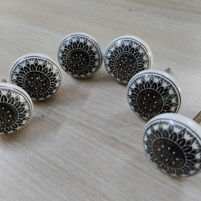 May include: Six white ceramic cabinet knobs with a black floral design. The knobs have a round shape and a metal screw for installation. The floral pattern is symmetrical and detailed, adding a decorative touch to furniture.
