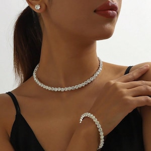 May include: A dazzling jewellery set comprising a necklace, bracelet, and stud earring. The necklace and bracelet are made up of many clear, round stones. The earring is a single, matching stone. The jewellery is set against a black background.