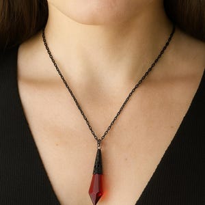Könnte beinhalten: Eine schwarze Halskette mit Anhänger. Der Anhänger hat einen facettierten, roten Edelstein in einer schwarzen, verzierten Metalleinfassung. Die Halskette wird über einem schwarzen Oberteil getragen.