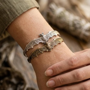 Op de afbeelding: Twee metalen armbanden met een vleermuisontwerp met gespreide vleugels. De ene armband heeft een zilverkleurige afwerking, terwijl de andere een bronskleurige afwerking heeft. Beide armbanden tonen gedetailleerde texturen op de vleugels.