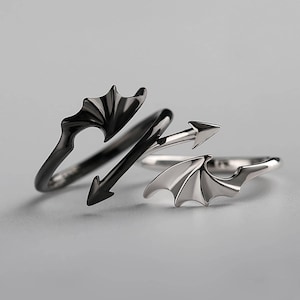 Może przedstawiać: Trzy pierścionki o tematyce fantasy. Jeden pierścionek jest czarny z motywem skrzydła nietoperza, drugi srebrny z motywem skrzydła nietoperza, a trzeci czarny z motywem strzały. Pierścionki są na szarym tle.