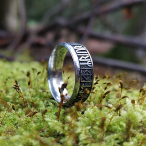 May include: A silver ring with engraved runic symbols. The ring is set against a backdrop of green moss and small plants. The ring's design features detailed, dark-filled engravings.