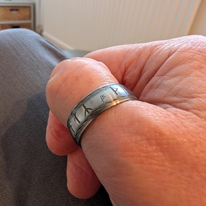 Może przedstawiać: Srebrny pierścień z wytrawionymi symbolami runicznymi. Pierścień jest noszony na palcu, prezentując szczegółowe grawerunki. Projekt zawiera opaskę z serią pionowych i ukośnych linii, tworzącą teksturowany wygląd. Metaliczny połysk pierścienia jest widoczny.