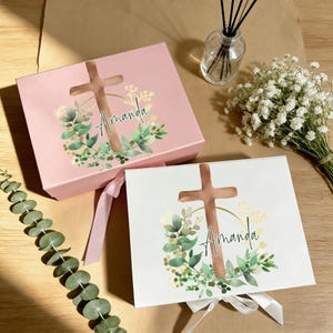 Può includere: Due scatole regalo, una rosa e una bianca, con un disegno a croce e il nome "Amanda" in corsivo. Le scatole sono decorate con una ghirlanda floreale e legate con nastri. Un mazzo di fiori bianchi e un diffusore sono sullo sfondo.