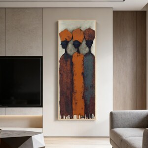 May include: Abstract artwork depicting three figures with orange headwear and dark profiles. The figures are rendered in hues of brown, orange, and blue-grey. The framed painting is displayed on a neutral wall, enhancing the modern aesthetic of the space.