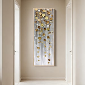 以下が含まれることがあります： 銀と金の抽象的なデザインの垂直ウォールアート。金色の花のような形が、銀色の背景に細い線から吊り下げられています。このアートワークは、明るい色の木製フレームに額装されています。